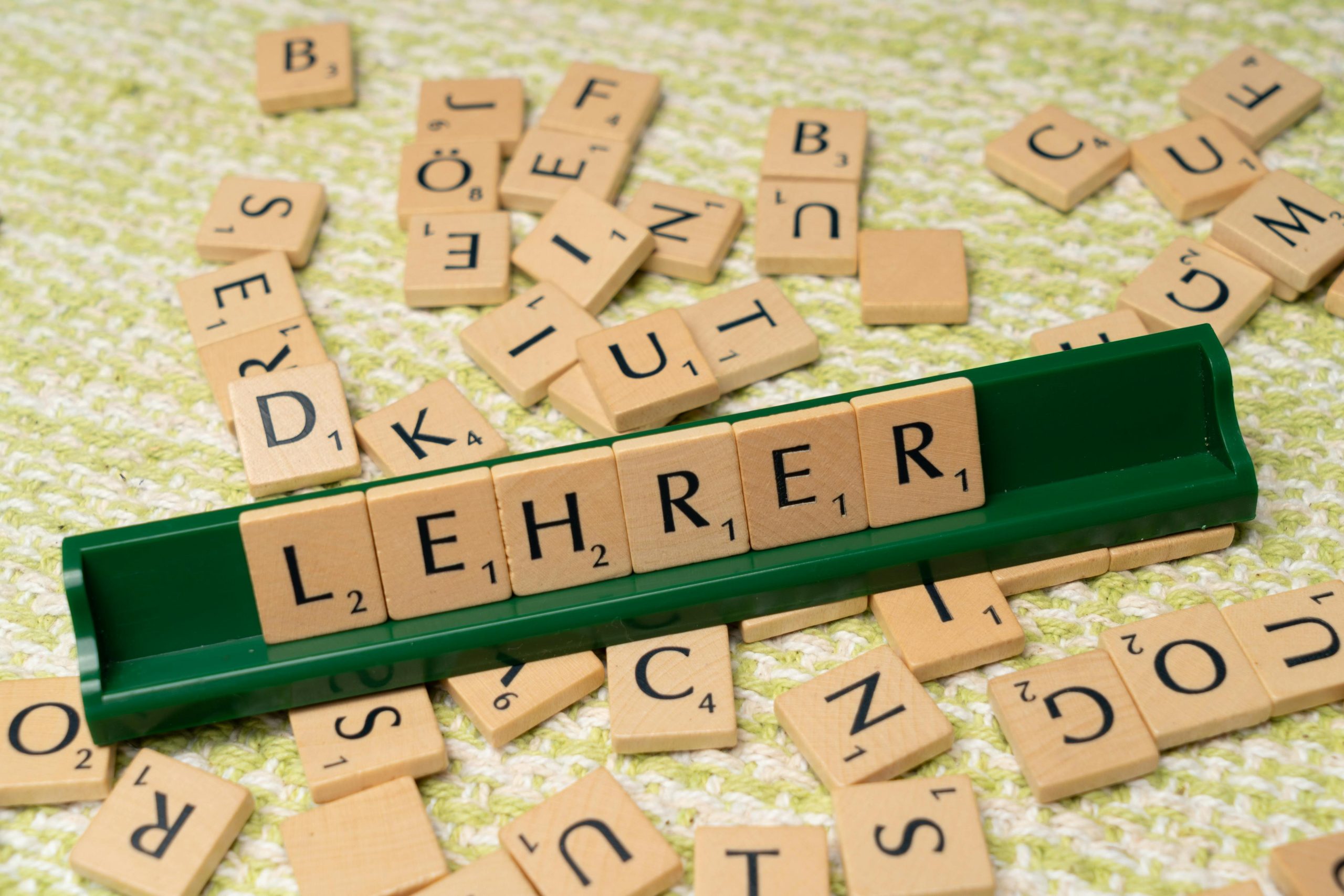 Über uns Wooden letter tiles spelling 'Lehrer' on a green rack, surrounded by scattered tiles.